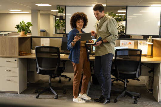 Reviewing Diverse coworkers standing at shared desk in open-plan office, with tablet and notebook