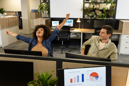 Diverse coworkers stretching and smiling in open plan office, with monitors showing graphs