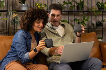 Sitting Diverse couple using silver laptop in living room on leather couch, holding dark gray mugs