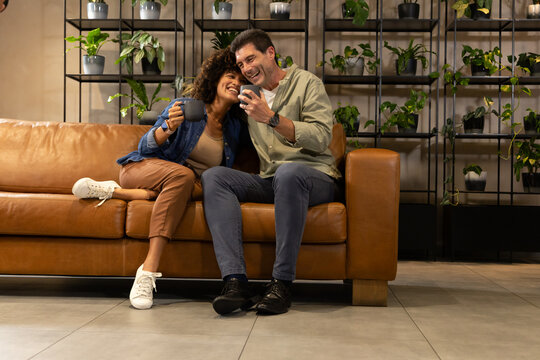 Smiling Diverse couple leaning on brown leather sofa in lounge, with smartphone and coffee mug