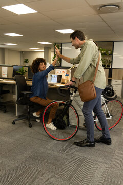 High-fiving diverse colleagues exchanging greetings in modern office, featuring bicycle and laptops