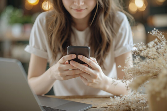 Customer using smartphone to contact online company's service team, holding the phone with both hands and typing. Blurry background shows a laptop and decorative elements