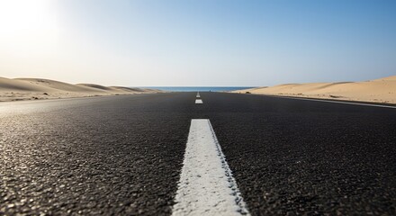 Fototapeta premium A long asphalt road stretches through a desert landscape towards the distant sea under a clear blue sky