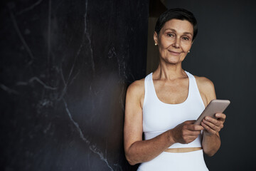 Portrait of middle aged Caucasian woman smiling while holding smartphone, standing against dark marble wall, wearing sportswear, looking directly at camera, short hair visible
