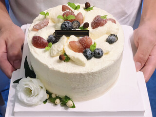 Two hands holding a white cake box with a decorated cream cake topped with grapes, blueberries, mint leaves, flower buds, and a blank card, surrounded by white flowers and greenery.