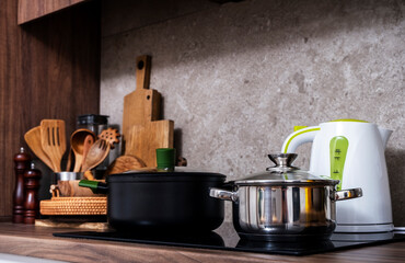 Steel pots on induction hob and kettle on cozy modern kitchen with cooking utensils, pots, cutting boards on wooden countertop