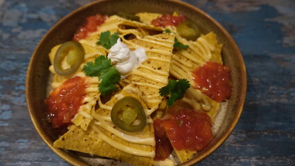 Close-up of loaded nachos topped with melted cheese, jalapeño slices, salsa, sour cream, and cilantro in a rustic bowl.
