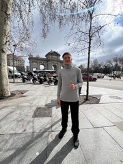 City Explorer's Stroll: A young person strolls confidently along a city sidewalk, their thumb raised in a gesture of optimism, with a majestic arch providing a stunning backdrop.