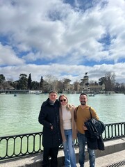 Friends by the Lake: Three friends pose happily near a serene lake with an ancient building as a backdrop, enjoying a leisurely day out under a vibrant blue sky with fluffy clouds.