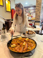 Exquisite Paella Presentation: A woman delicately serves a vibrant paella dish, featuring succulent shrimp and zesty lemon wedges, within a classic dining establishment.