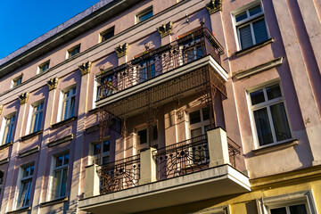 Facade of a historic townhouse with pink plaster, decorative cornices, and a wrought iron balcony, illuminated by afternoon sunlight against a clear blue sky.