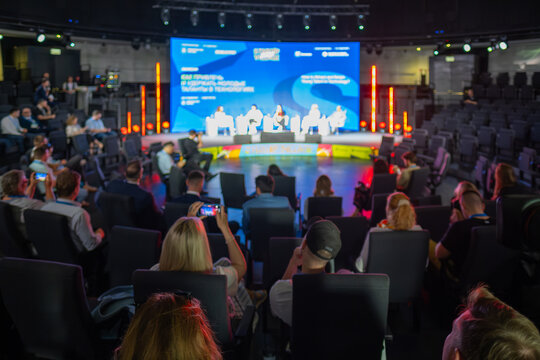 Conference attendees engaging in discussion, watching speakers present on stage under focused lighting.