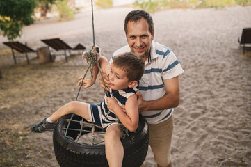 dad swinging little son on tire swing in park in summer on beach. Outdoor games with child in summer. father and son spending time together