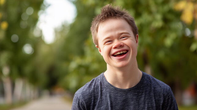 Young smiling man with special needs laughing outdoors, trees in the background. t-shirt. Close-up portrait of a happy young male teenager with Down syndrome. Acceptance of diversity, body inclusive.