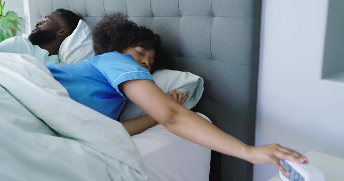 Diverse couple waking by alarm clock, woman switching off alarm as partner stretching in dawn light