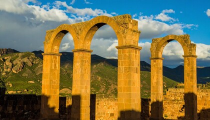 Sunlit ancient stone arches standing tall against mountainous landscape and dramatic sky.