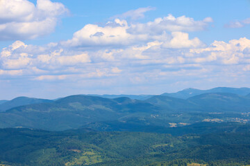 High mountains covered with dense forests are sinking into a blue haze. In the background, there's an endless sky with big white clouds. This is a wonderful summer landscape, full of tranquility and n