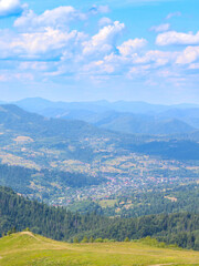 A high-angle view of a picturesque valley with a village, surrounded by wooded mountains. Summer green slopes in the foreground, a blue haze in the background, and white clouds in the sky create a sen © Олег Струс