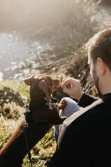 Man in black long sleeved shirt and blue jeans sitting out in nature next to a lake with a brown dachshund in a harness and on leash engaging with his pet dog. Sun flare. Lake and grass