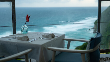 Romantic oceanfront dining table with a single red rose in a vase, set with napkins and cutlery, overlooking turquoise waves and a scenic coastal view.