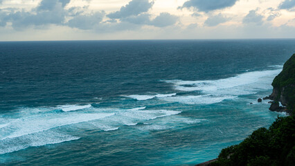 A breathtaking view of the turquoise ocean waves crashing against a lush green cliffside under a cloudy sky, capturing the serene beauty of nature’s power and tranquility.