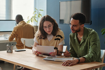Young woman reading document and discussing it with her colleague while they working in team at table at office