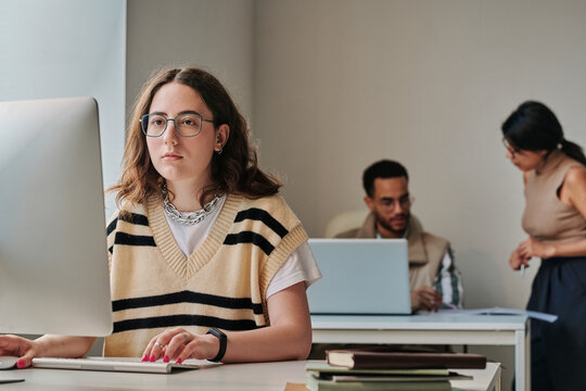 Young office worker in eyeglasses sitting at her workplace in front of computer monitor and working on computer with colleagues in background