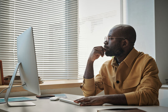 African young businessman in eyeglasses looking at window while thinking over new ideas at his workplace with computer monitor