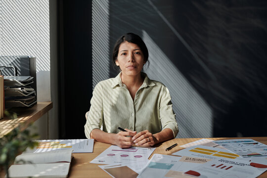Portrait of young manager looking at camera while working with documents at her workplace at office