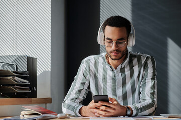 Young businessman in wireless headphones using smartphone for online communication with his colleague at table at office