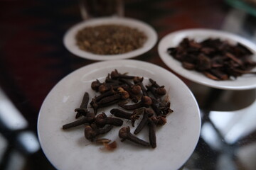 Aromatic dried cloves displayed on a small white plate, with other spices blurred in the background on a reflective surface.
