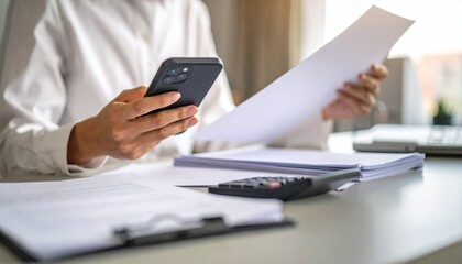 Fototapeta premium A person flipping through documents on a desk and checking their smartphone