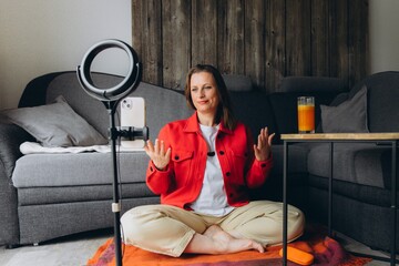Smiling woman filming video with smartphone and ring light at home. Sitting cross-legged on the floor, she creates content, talks to camera, remote online communication or vlogging session