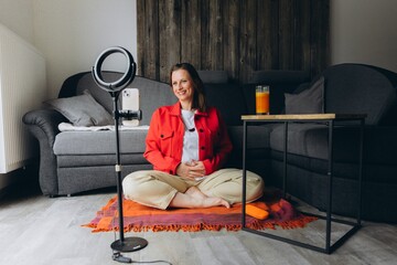 Smiling woman filming video with smartphone and ring light at home. Sitting cross-legged on the floor, she creates content, talks to camera, remote online communication or vlogging session