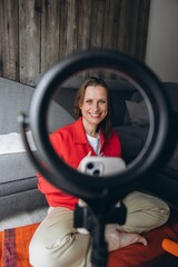 Smiling woman filming video with smartphone and ring light at home. Sitting cross-legged on the floor, she creates content, talks to camera, remote online communication or vlogging session