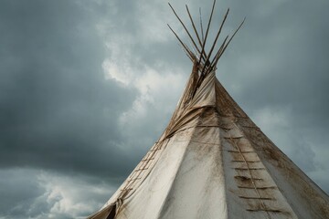 Low-angle view of a traditional tipi under a dramatic sky.  The tipi's pale, slightly weathered canvas is supported by numerous wooden poles, which radiate outward from the apex.  