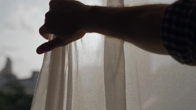 Male hand opens window curtain or drape to reveal sunset or sunrise outdoors. Man moves fabric to show the light of day outside in city or urban view. Warm sunlight shining in creating artistic flare