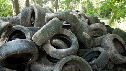A large stack of dirty, used tires sits in a forest clearing. The tires are scattered and abandoned among the trees. This scene raises awareness about waste management and recycling. © bisonov