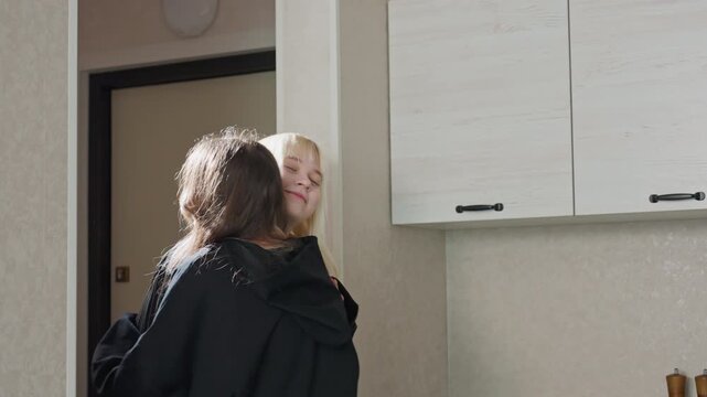 woman hugging sister upon return from journey while sister walks away after she drops handbag on chair then retrieves cheese snack from bag and places it on table in warm cozy kitchen setting