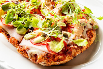 Close-up of gourmet flatbread with tomatoes, ham, melted cheese, arugula, green sauce, and dressing on a crispy crust, served on a white plate