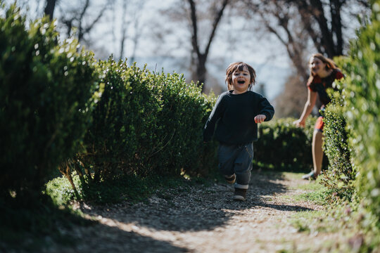 A joyful child runs along an outdoor pathway surrounded by lush green hedges while a parent follows in playful pursuit under sunlight, embodying happiness, outdoor activity, and family bonding.