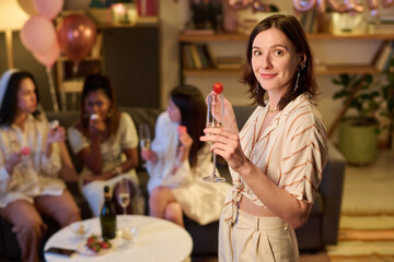 Portrait of young Caucasian woman smiling and holding champagne glass with strawberry garnish, standing in foreground while diverse group of young women socializing on sofa at bachelorette hen party