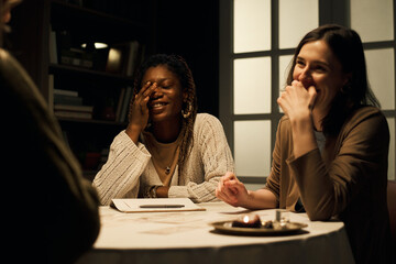 Young Black and Caucasian girlfriends laughing together during tarot card reading, sitting at...