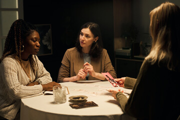 Fototapeta premium Three young women, Black and Caucasian, sitting around round table, engaged in mystic tarot reading session, discussing boyfriend on photograph, while consulting psychic for relationship advice