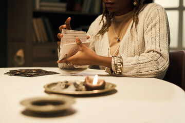 Black woman shuffling tarot cards at round table with burning candle in foreground, wearing jewelry and sweater, partial face visible, appearing as young adult