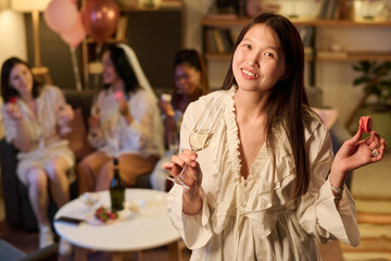 Portrait of young Asian woman smiling at camera, holding glass of wine and macaron, with three young adult women sitting in background, celebrating bridal or engagement party indoors
