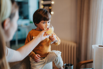 A small boy being helped by a parent to drink juice from a glass while sitting indoors. The scene exudes comfort, care, and warmth in a home environment.