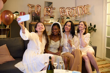 Group of four young women, including Black and Caucasian, sitting on sofa taking selfie together at bridal shower celebration, holding champagne glasses, smiling at camera