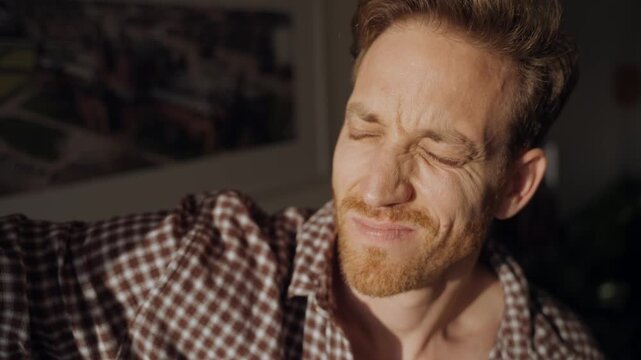 Man opens window blinds and shields eyes from bright sunlight, squinting and raising hand to block glare indoors. Morning light floods the room, creating strong shadows on his face. guy blinded by sun