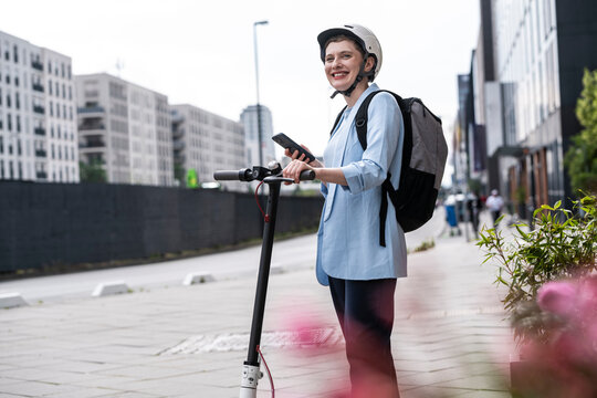 Woman with e scooter smiling outdoors in city with helmet and backpack